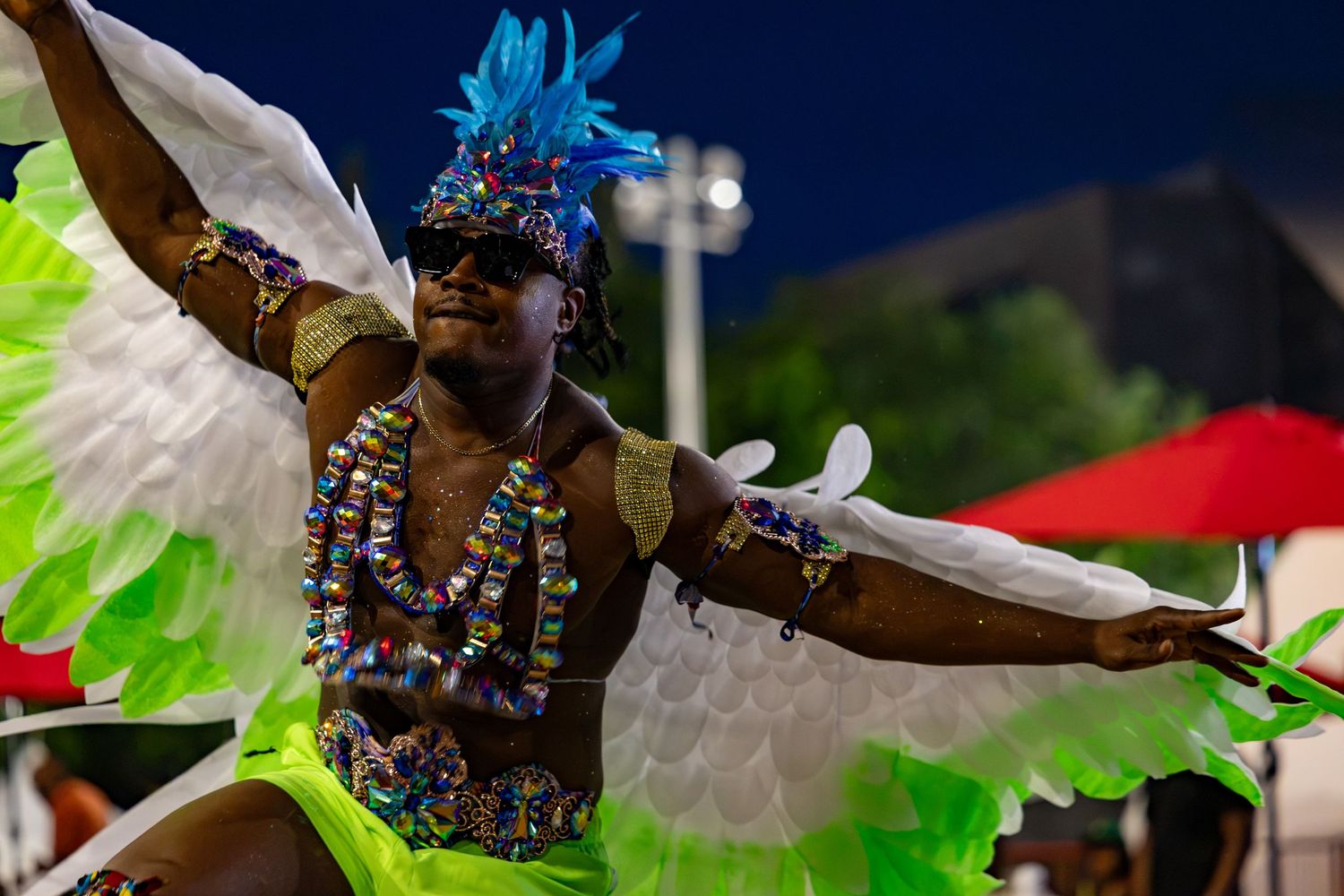 Colorful carnival dancer in green feathered costume and blue headdress performing at a night festival.