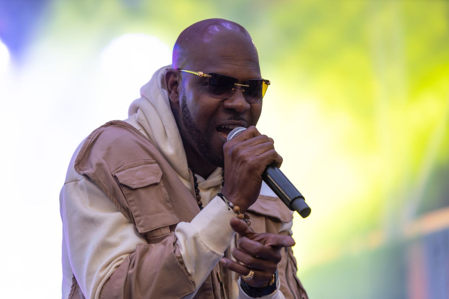 Bald male performer sings into microphone wearing beige hoodie and sunglasses on stage.