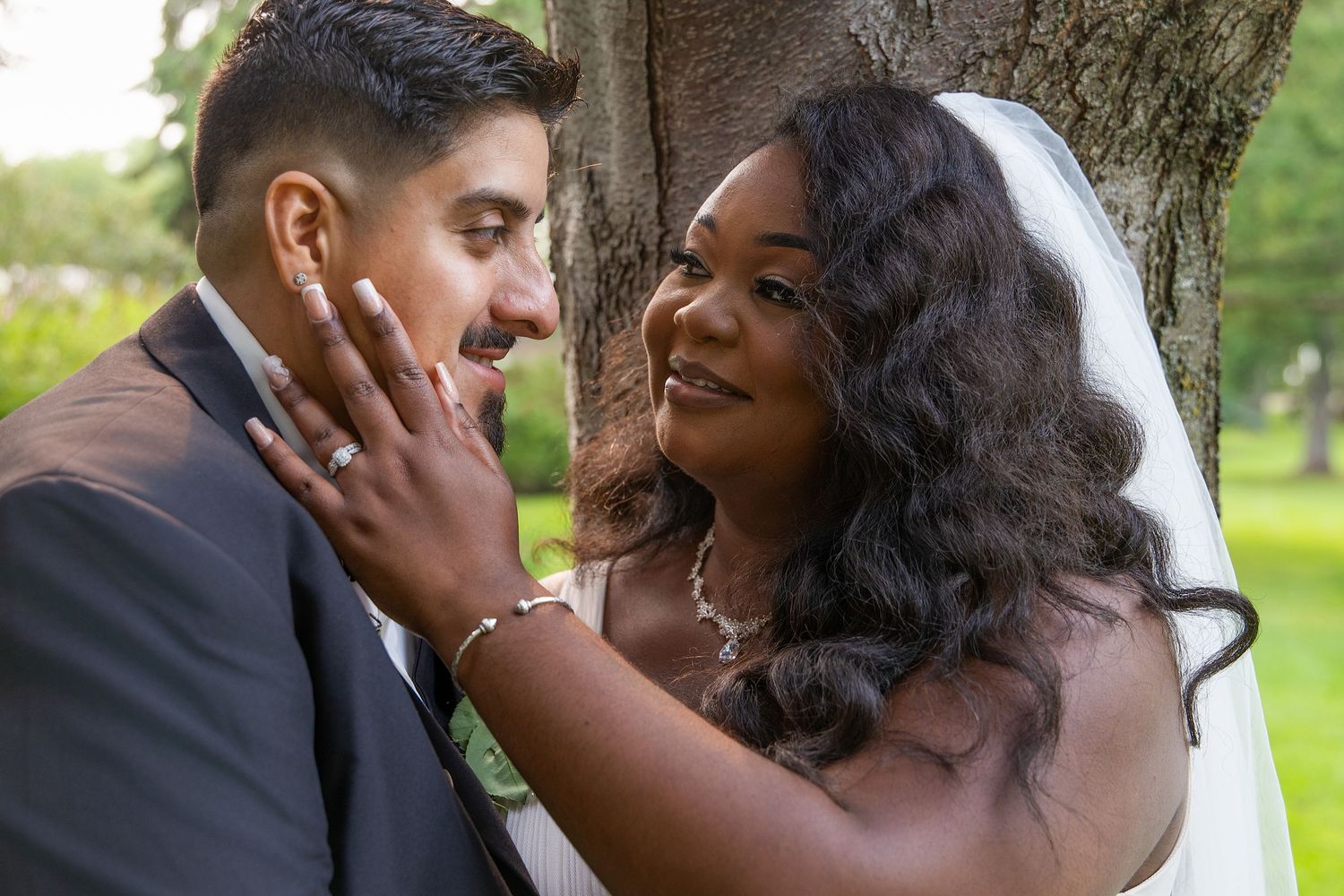 Interracial couple in wedding attire smiling intimately near a tree, bride in veil touching groom's face.