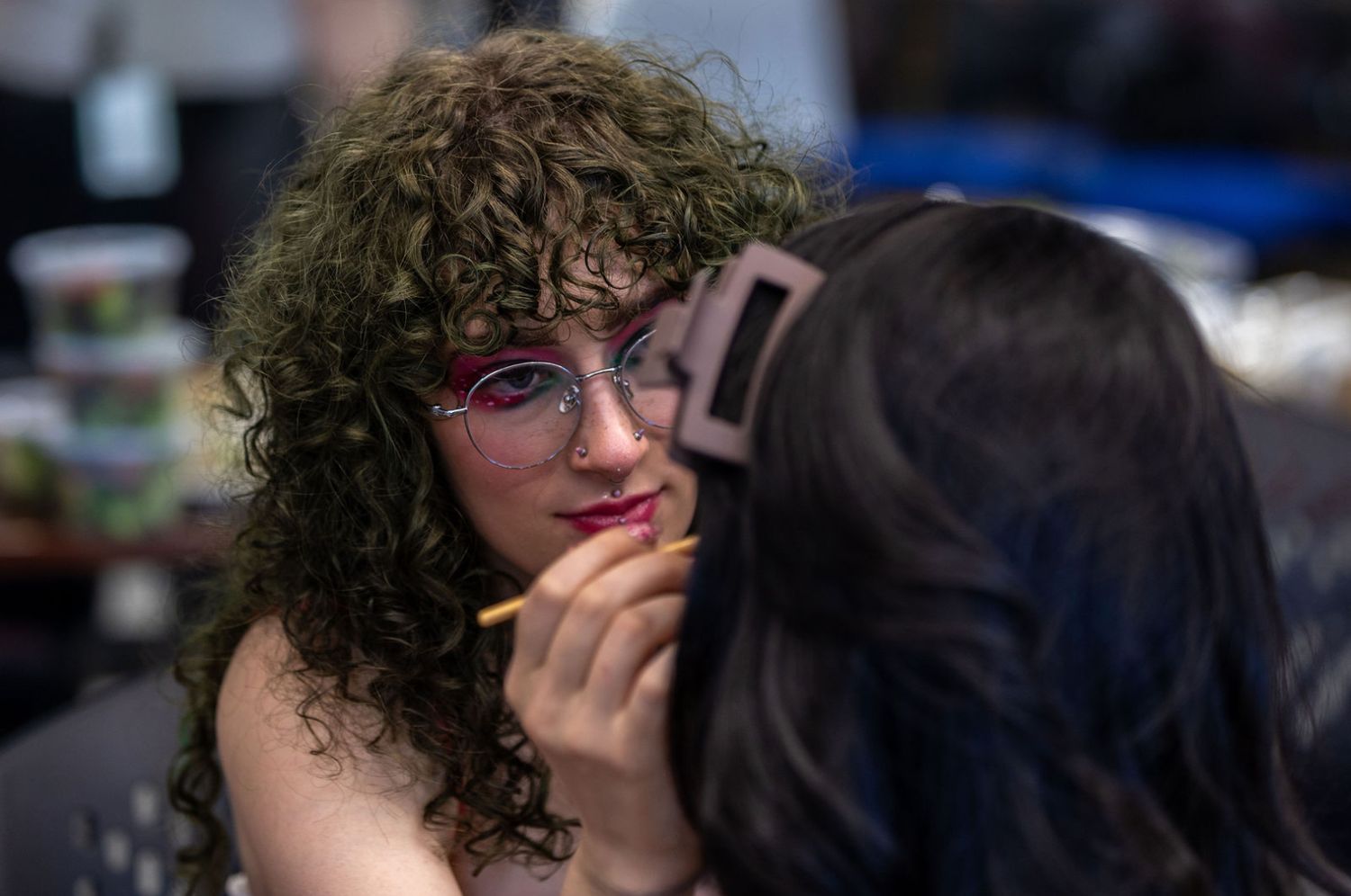 Young woman with curly hair and glasses getting lipstick applied by makeup artist indoors.
