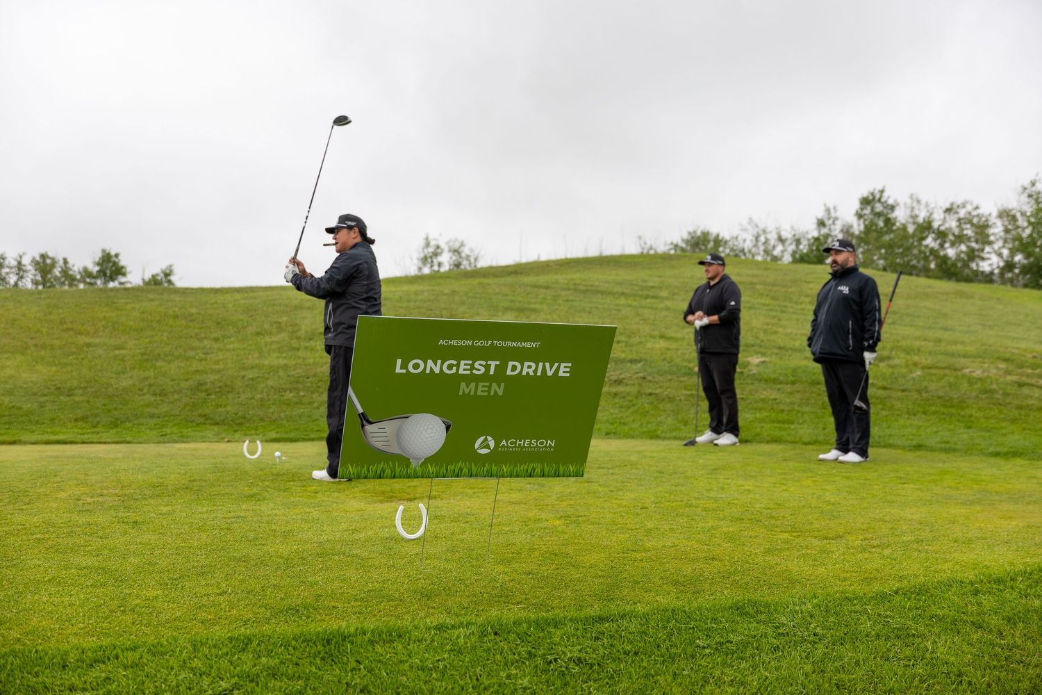 Golfer swinging a club at a Longest Drive Men competition on a lush green golf course with spectators watching.