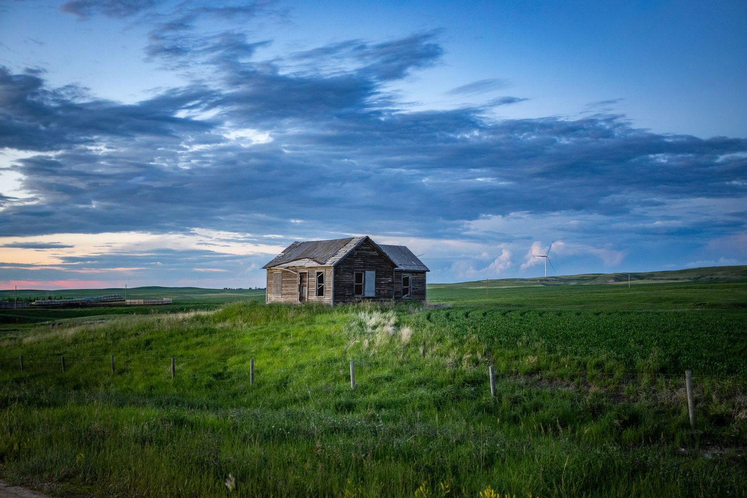 Abandoned stone farmhouse stands alone in vast green prairie under dramatic blue cloudy sky at dusk.