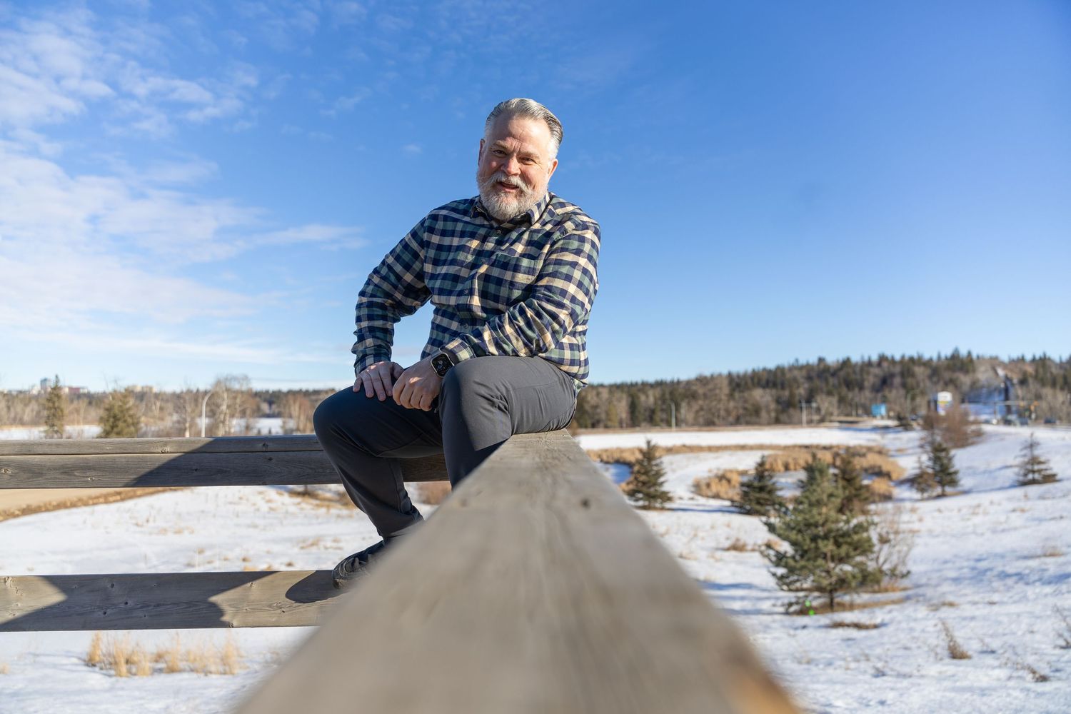 Bearded man in plaid shirt sits on wooden railing in snowy winter landscape under blue sky.