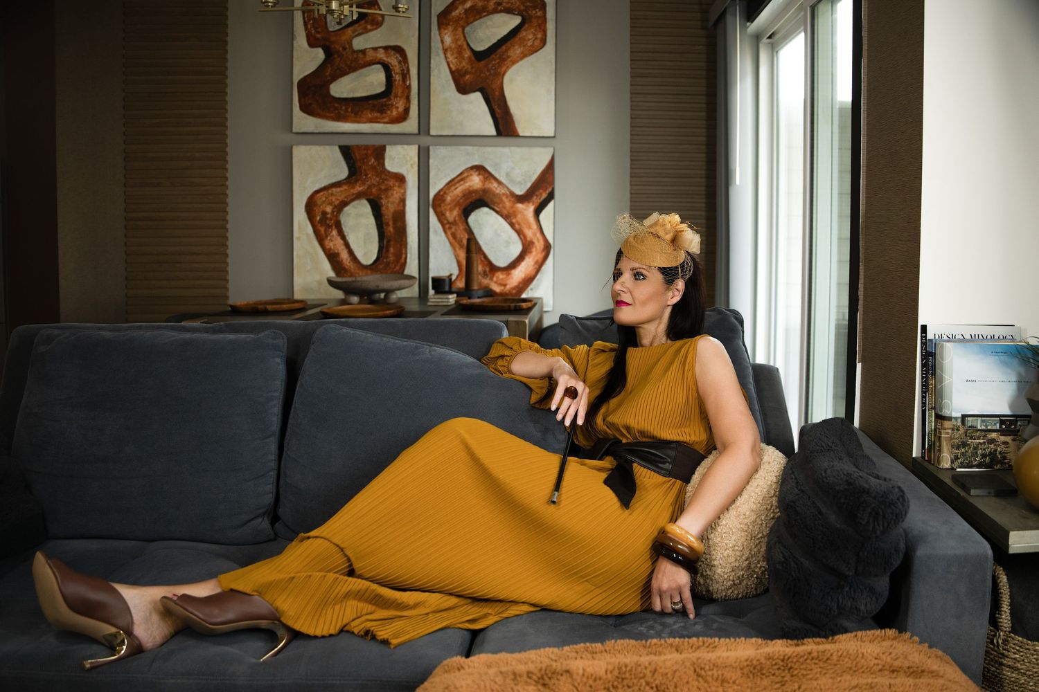 Woman in mustard yellow dress with black belt reclines on gray sofa, wearing gold headpiece in modern living room.
