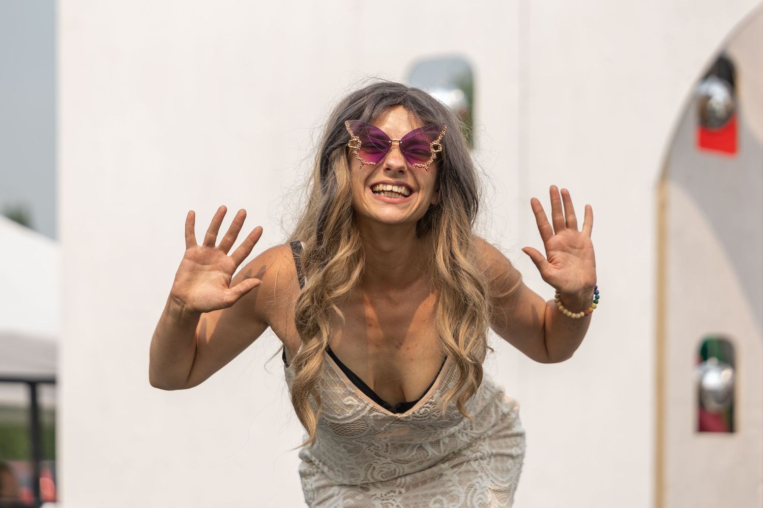 Laughing woman with heart-shaped pink sunglasses and wavy hair posing playfully with hands raised outdoors.