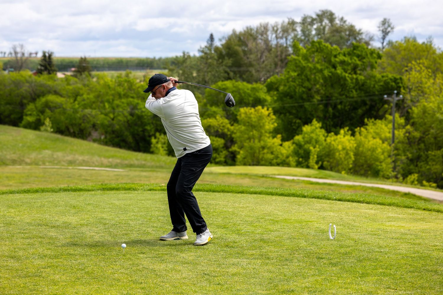 Golfer completing a full swing on a lush green golf course surrounded by trees on a cloudy day.
