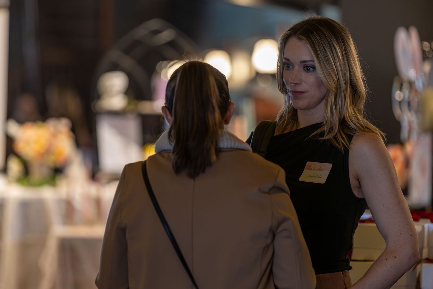 Two women networking at an indoor event, one wearing a black dress with a name tag.