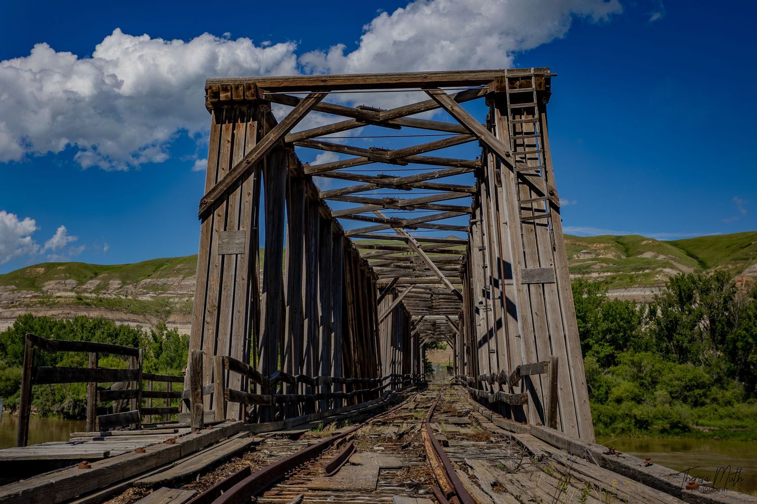 Rustic wooden railroad trestle bridge with steel truss framing stretches over a river under a blue sky.