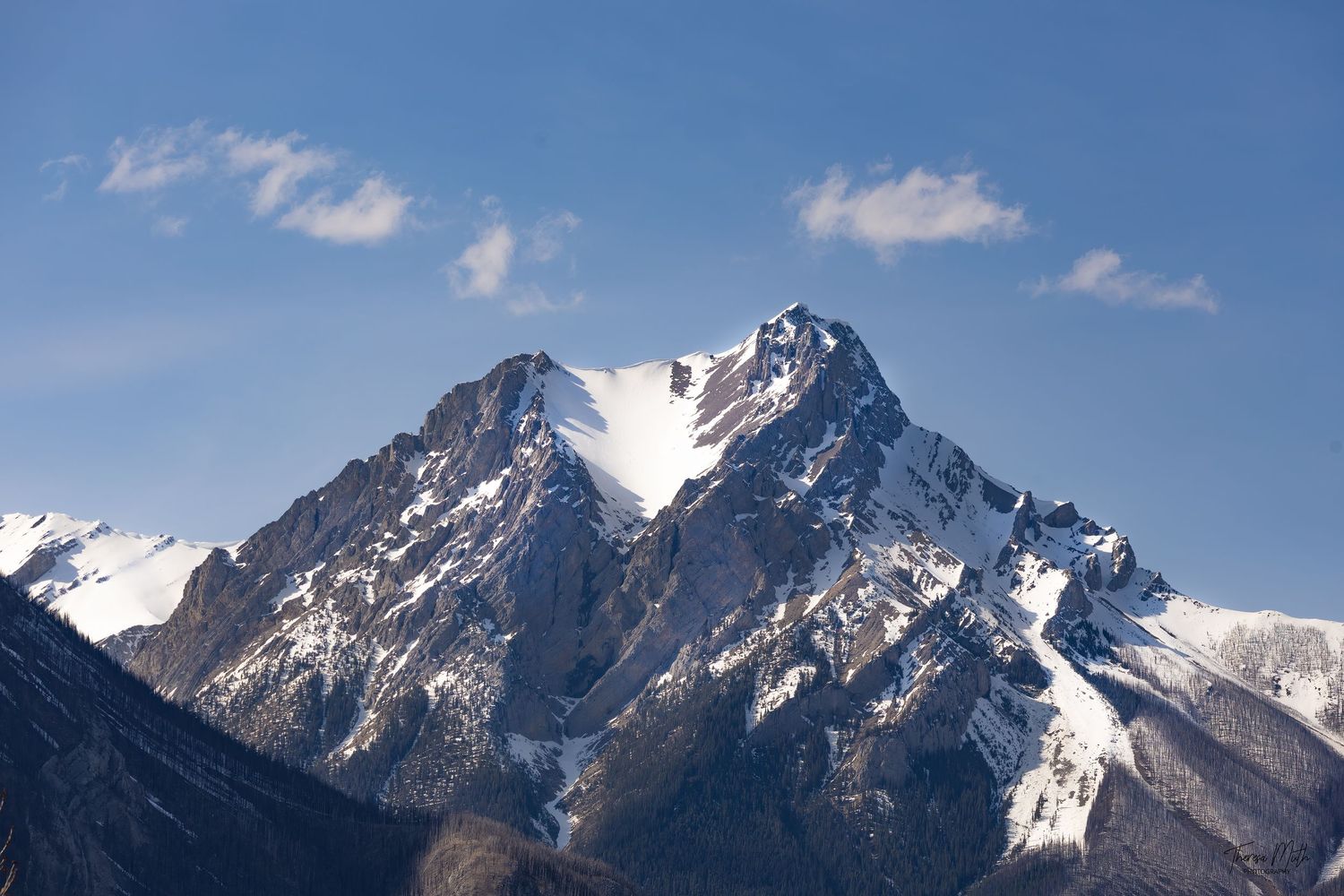 Snow-capped mountain peak rising under a clear blue sky with small white clouds in alpine setting.