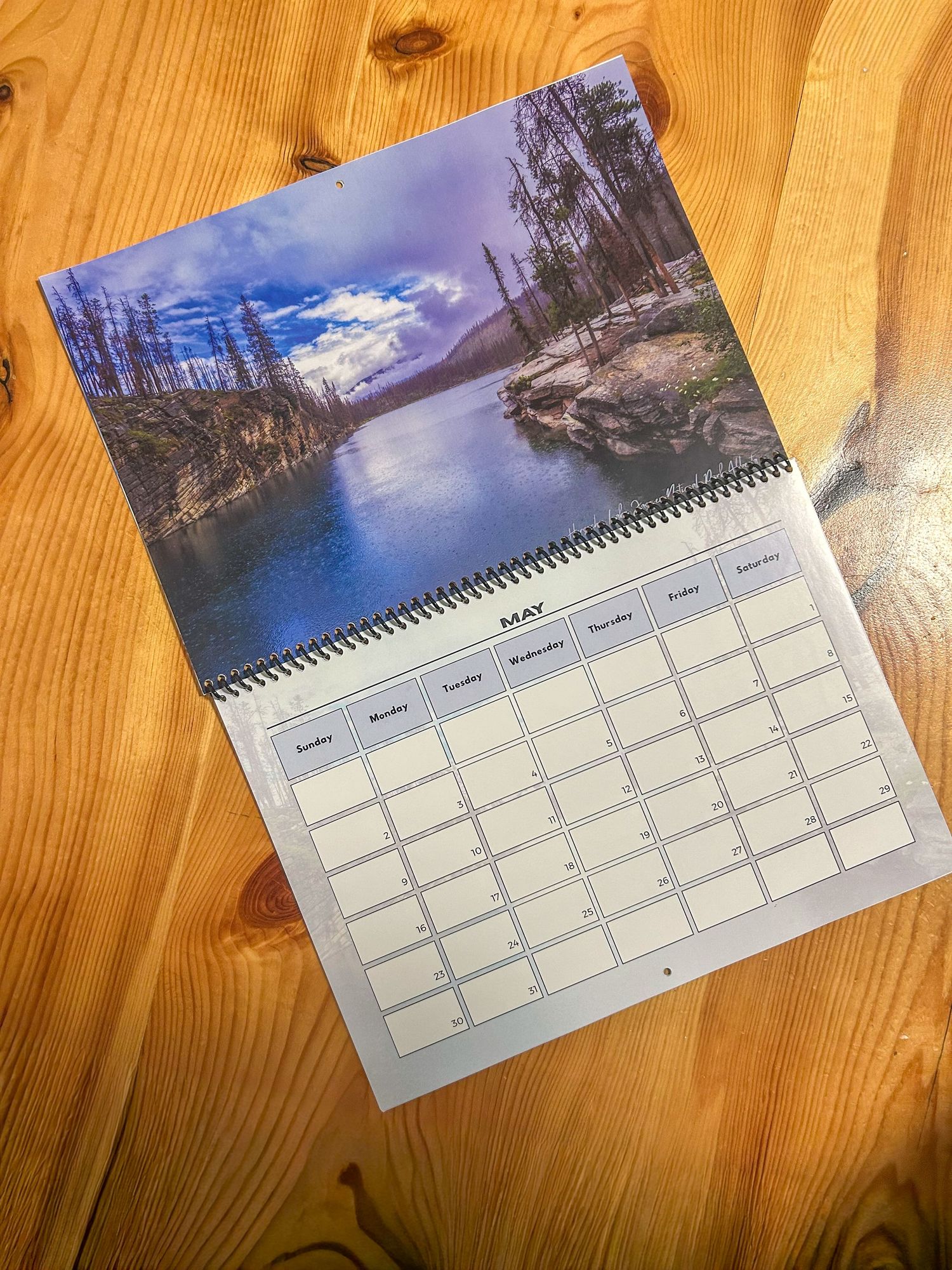 Spiral-bound wall calendar open to May featuring a scenic mountain river landscape photo, placed on a wooden surface.