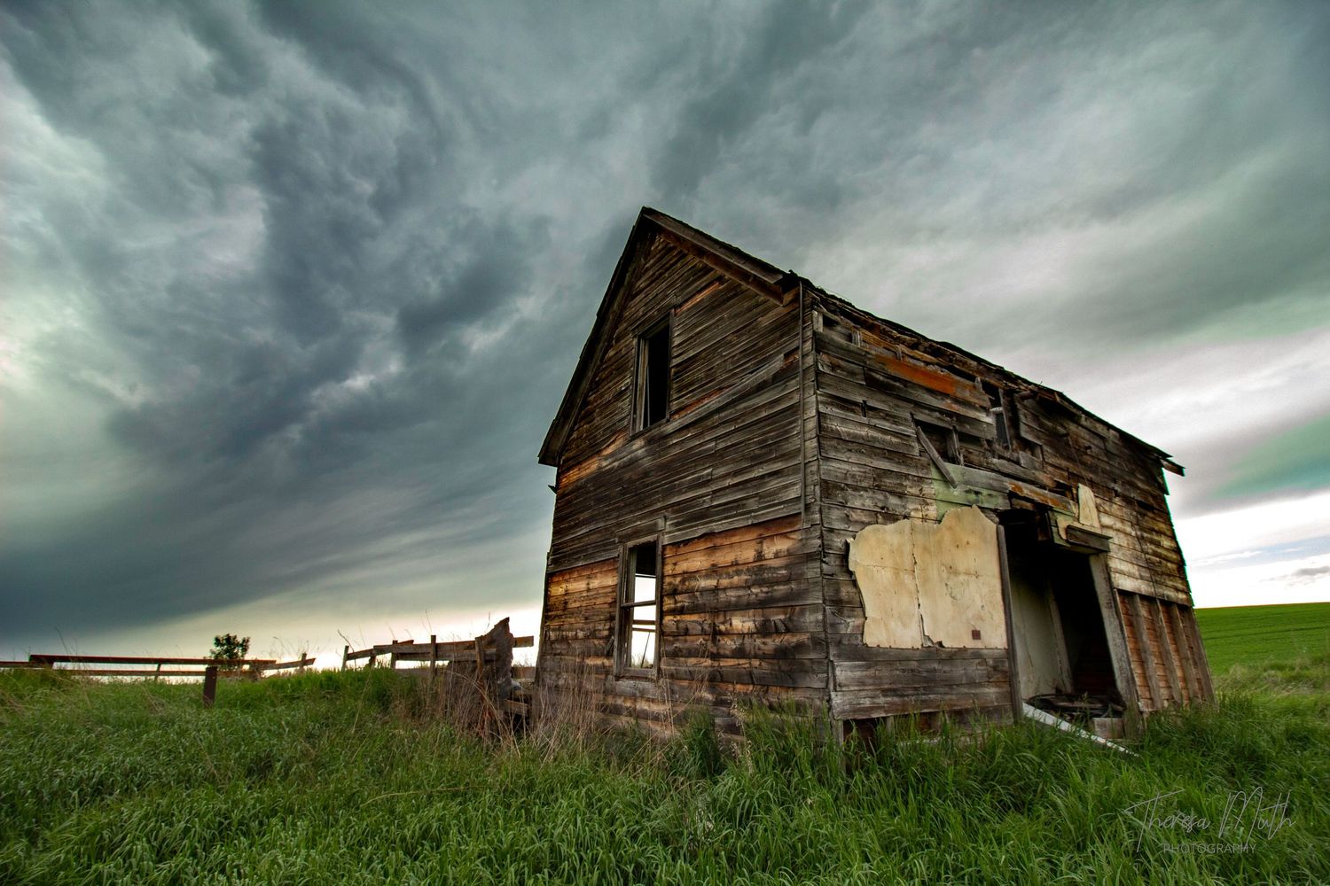 Abandoned wooden barn under dramatic stormy sky on green prairie landscape.