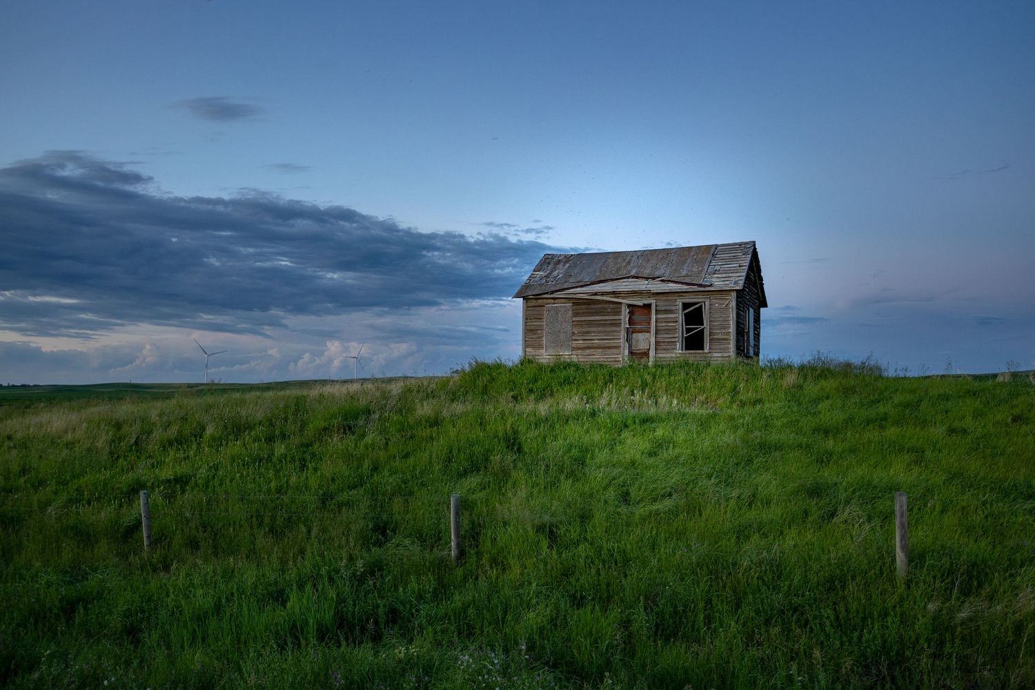 Abandoned wooden farmhouse on a green grassy hill under a dramatic blue cloudy sky at dusk.