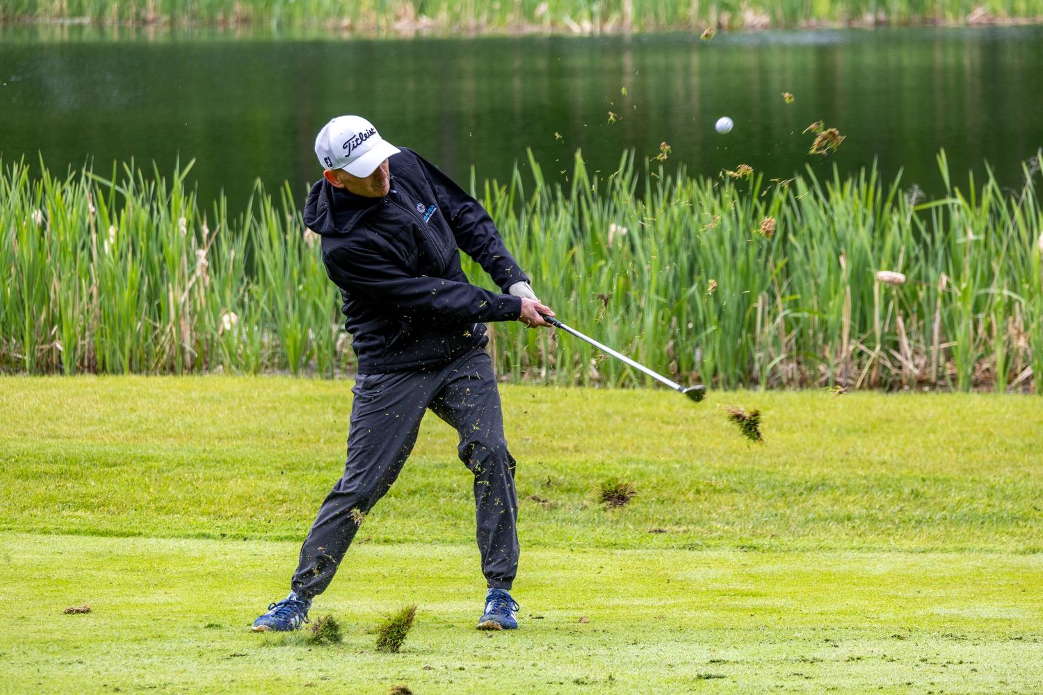 Golfer in black rain gear swinging a club near a pond on a lush green golf course.