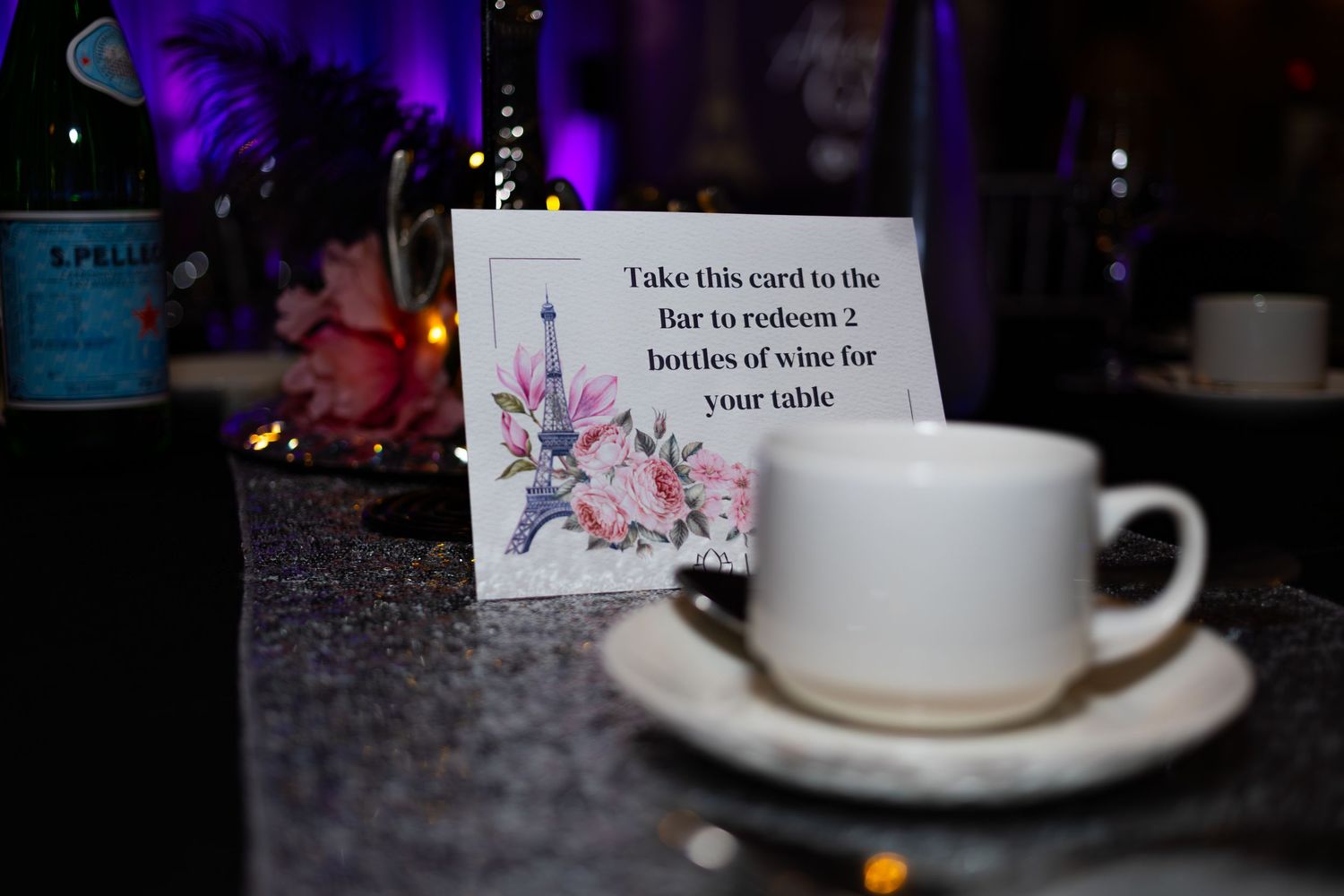 White card offering two free wine bottles at the bar displayed beside a teacup at a Paris-themed gala.