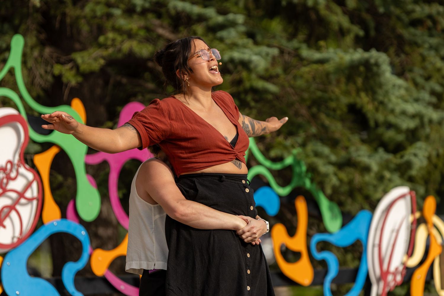 A joyful woman in a rust crop top dances outdoors with arms spread wide before a colorful abstract mural.