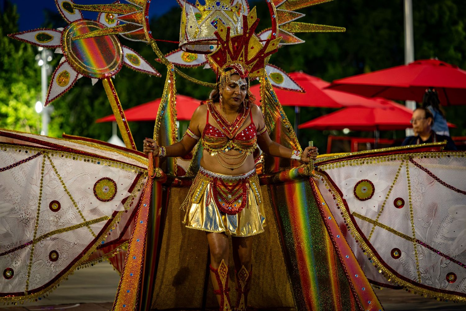 Vibrant carnival dancer in colorful gold and rainbow costume with large decorative wings performs at a festive night parade.
