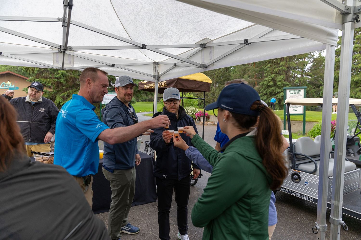 Group of golfers gathering under a white canopy tent at an outdoor golf event on a cloudy day.
