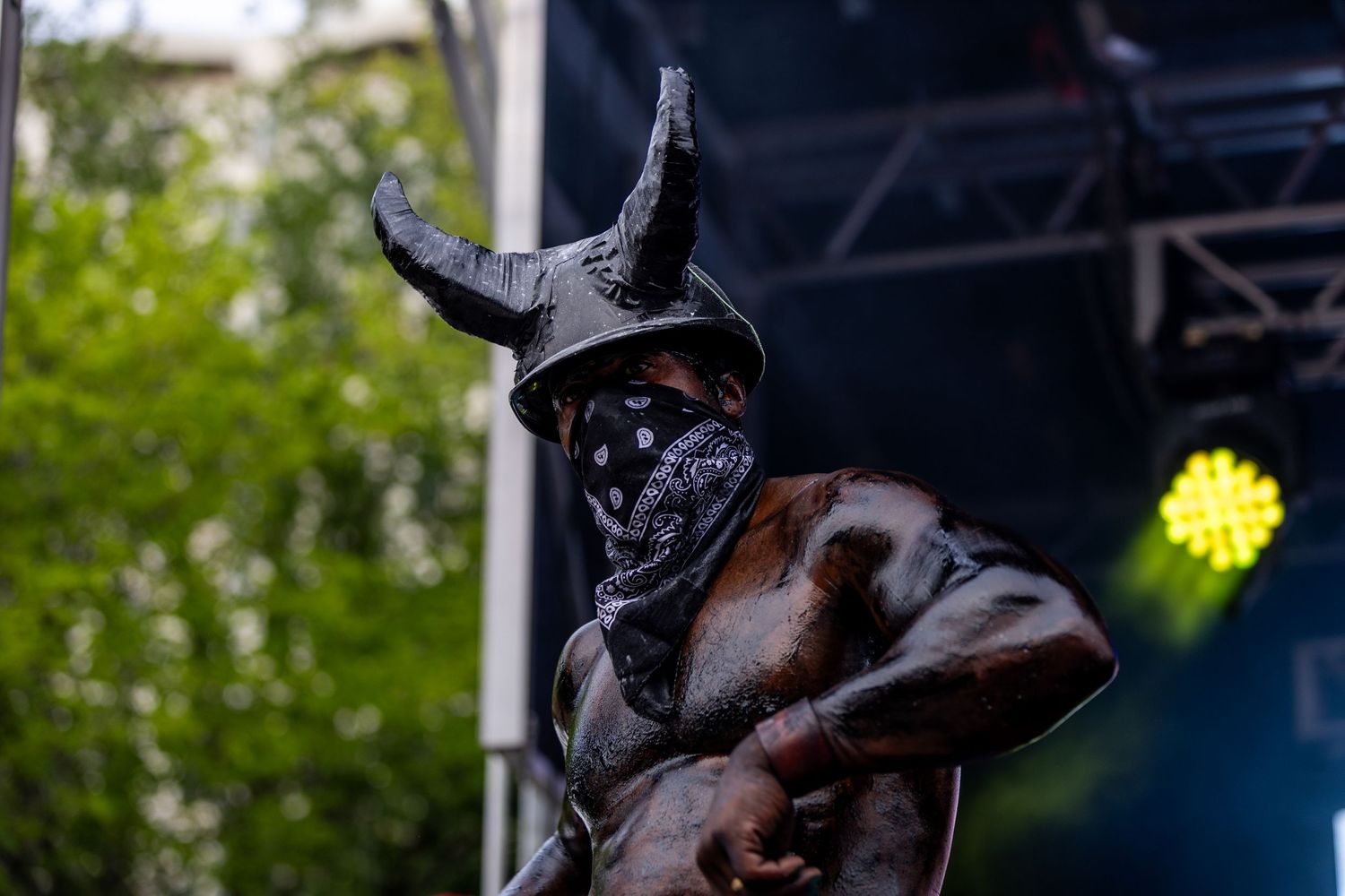 Mysterious performer wearing horned hat and face mask on outdoor festival stage with green trees.
