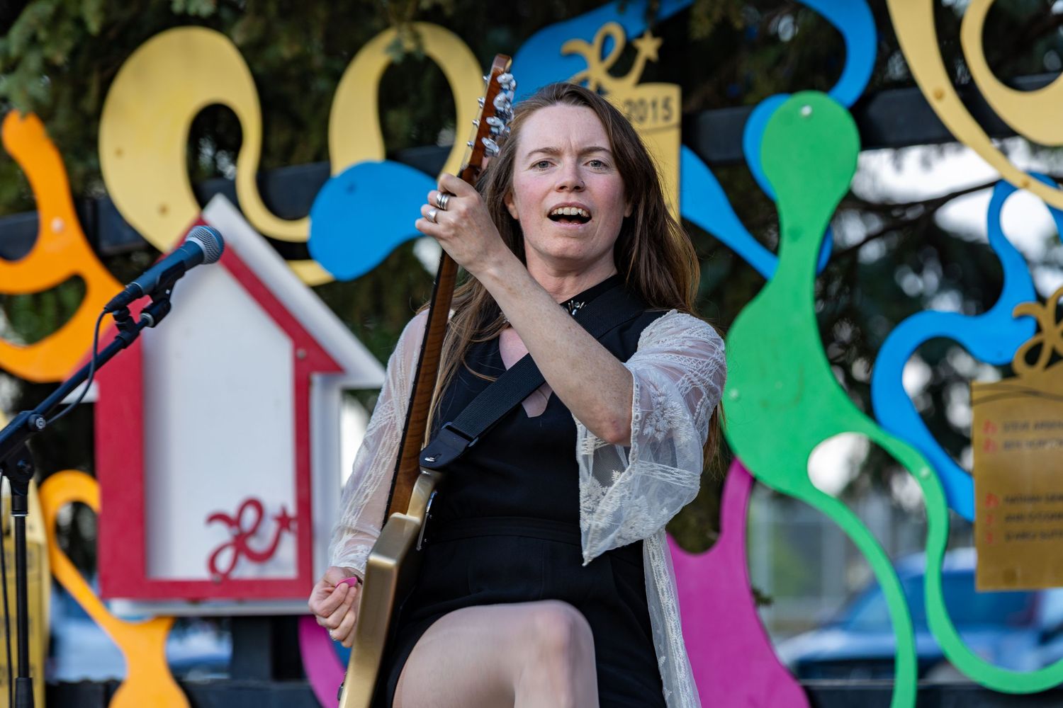 A female musician passionately plays electric guitar on an outdoor stage before a colorful art installation.