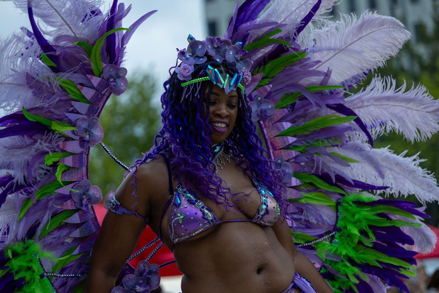 Woman in vibrant purple and green feathered carnival costume smiling during a colorful festival parade.
