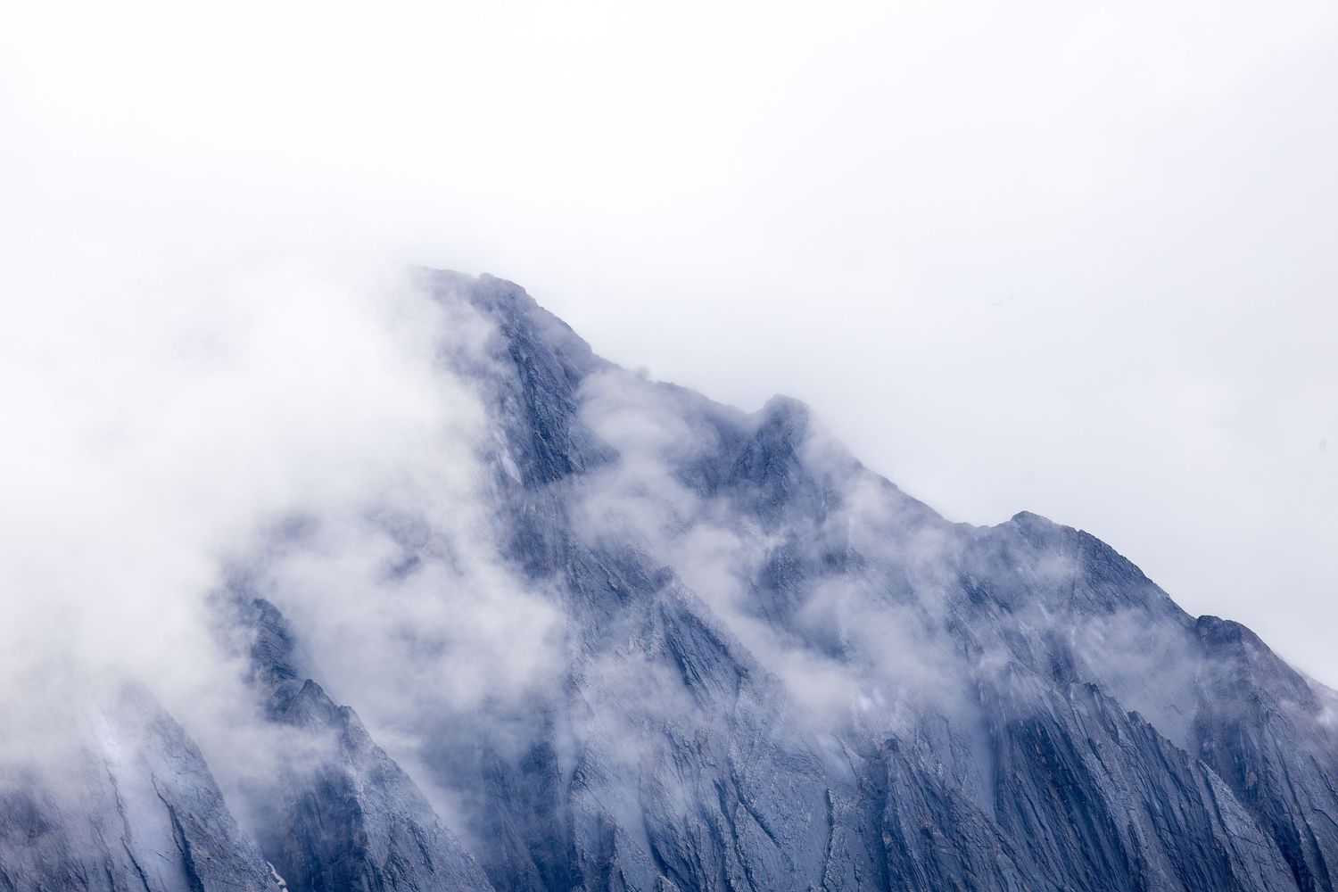 Steep rocky mountain summit dramatically enveloped in swirling white clouds and fog against pale sky.