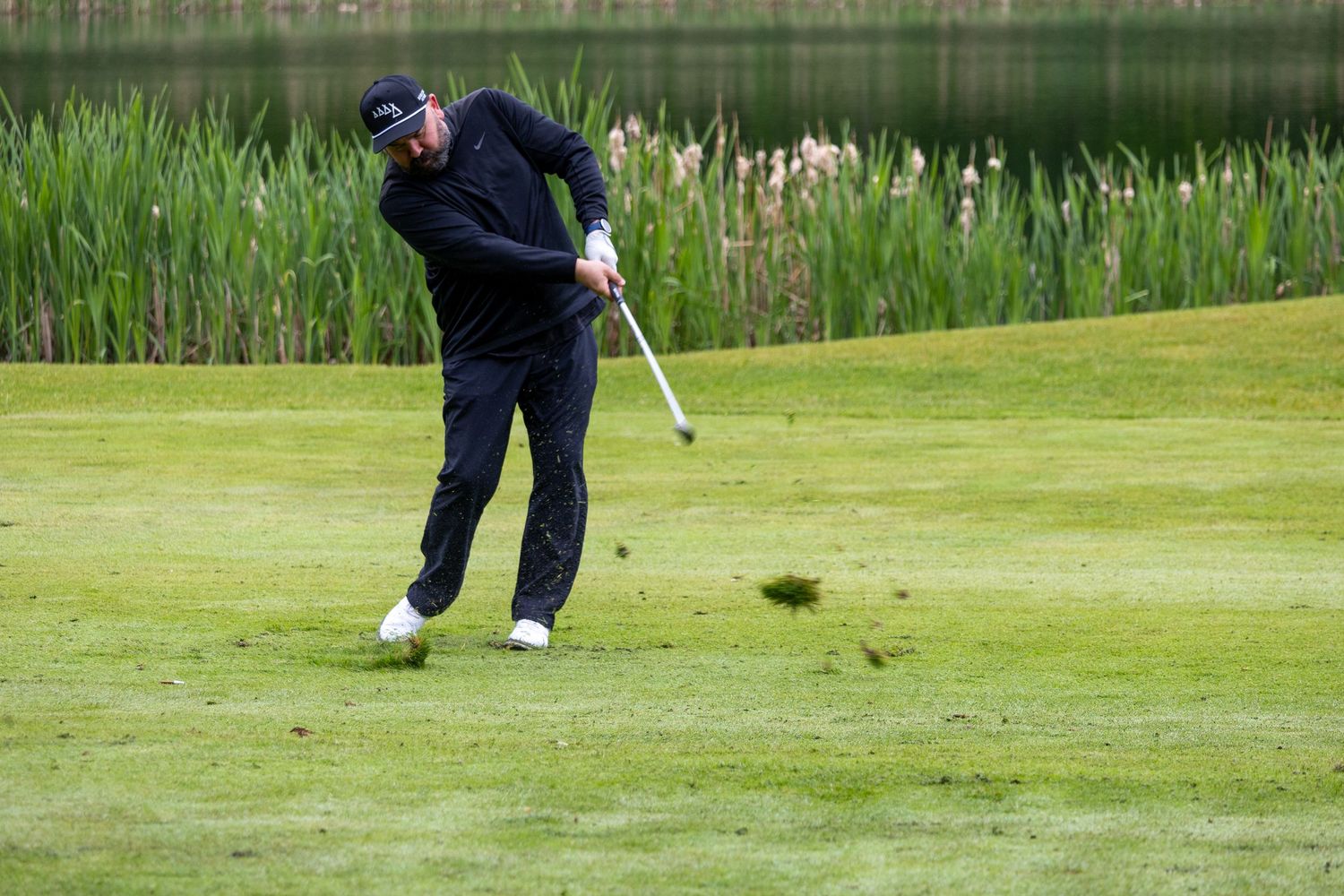 Golfer in black outfit swinging club on lush green fairway beside a tranquil pond.