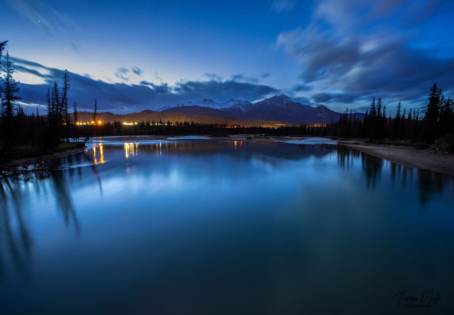 Blue hour twilight over a calm river reflecting city lights and silhouetted trees in a mountain valley.
