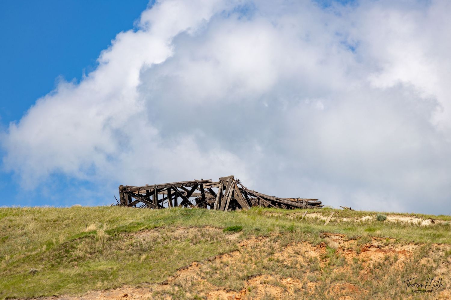 Collapsed wooden structure ruins on a grassy hillside under dramatic blue sky with white clouds.