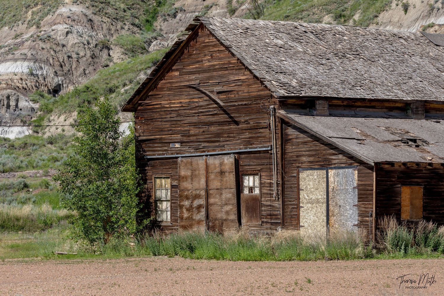 Abandoned weathered wooden barn building with boarded windows set against a rocky hillside and green trees.