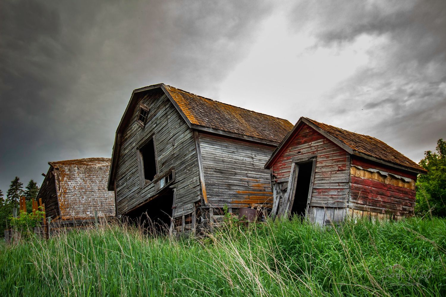 Abandoned weathered wooden barn leaning under dramatic stormy sky in rural countryside setting.