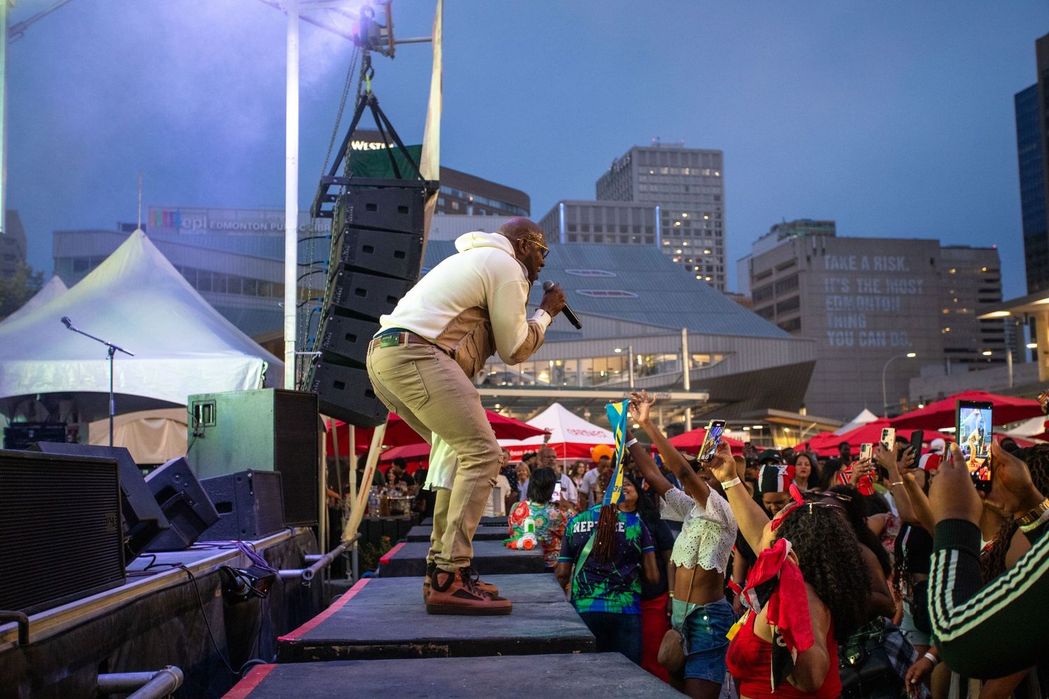 Male performer leans toward crowd on outdoor stage at dusk during urban music festival.