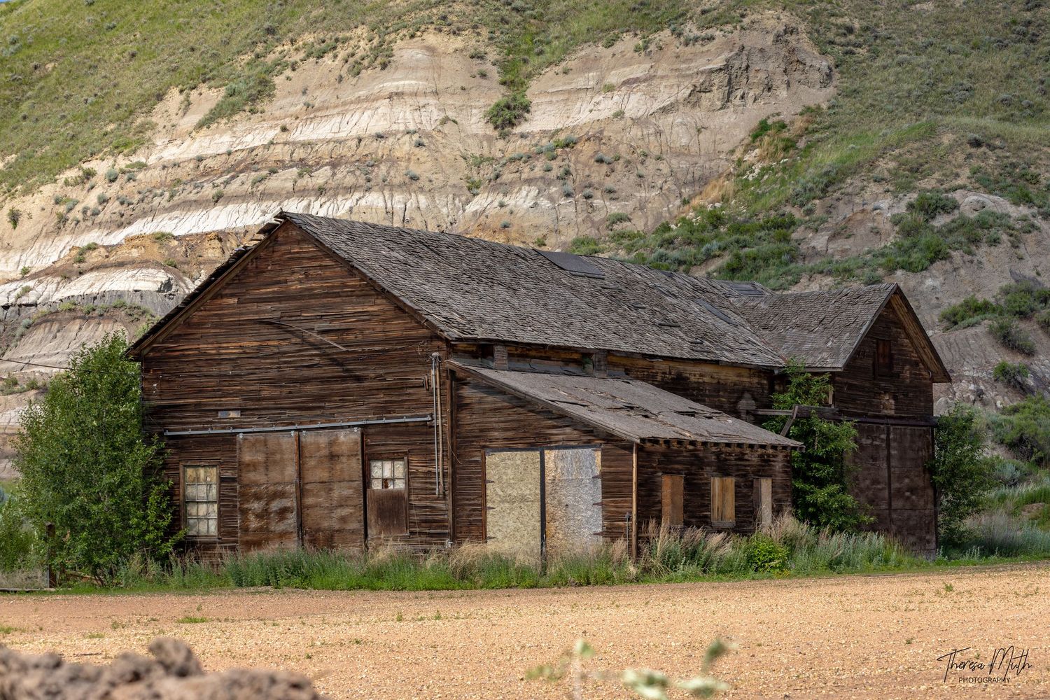 Abandoned weathered wooden barn stands against eroded badlands cliffs in a remote arid landscape.