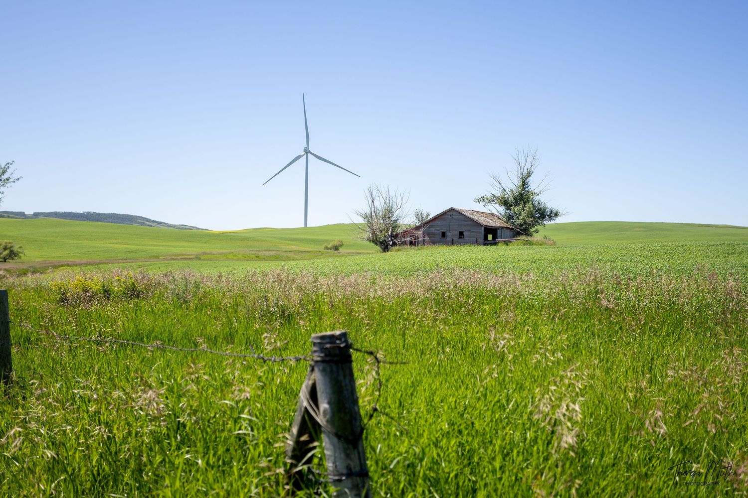 Green rolling hills with a wind turbine and rustic barn under a clear blue sky, framed by a wooden fence post.