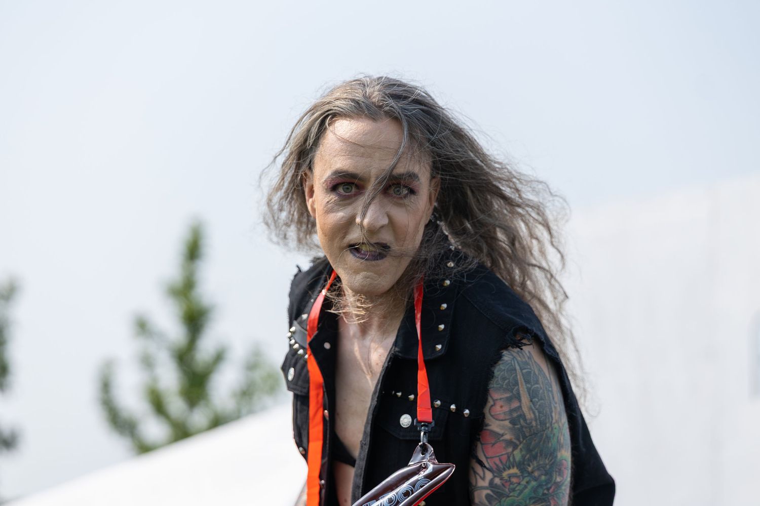 Older woman with long gray hair, tattoos, and a black vest wearing a red lanyard outdoors.