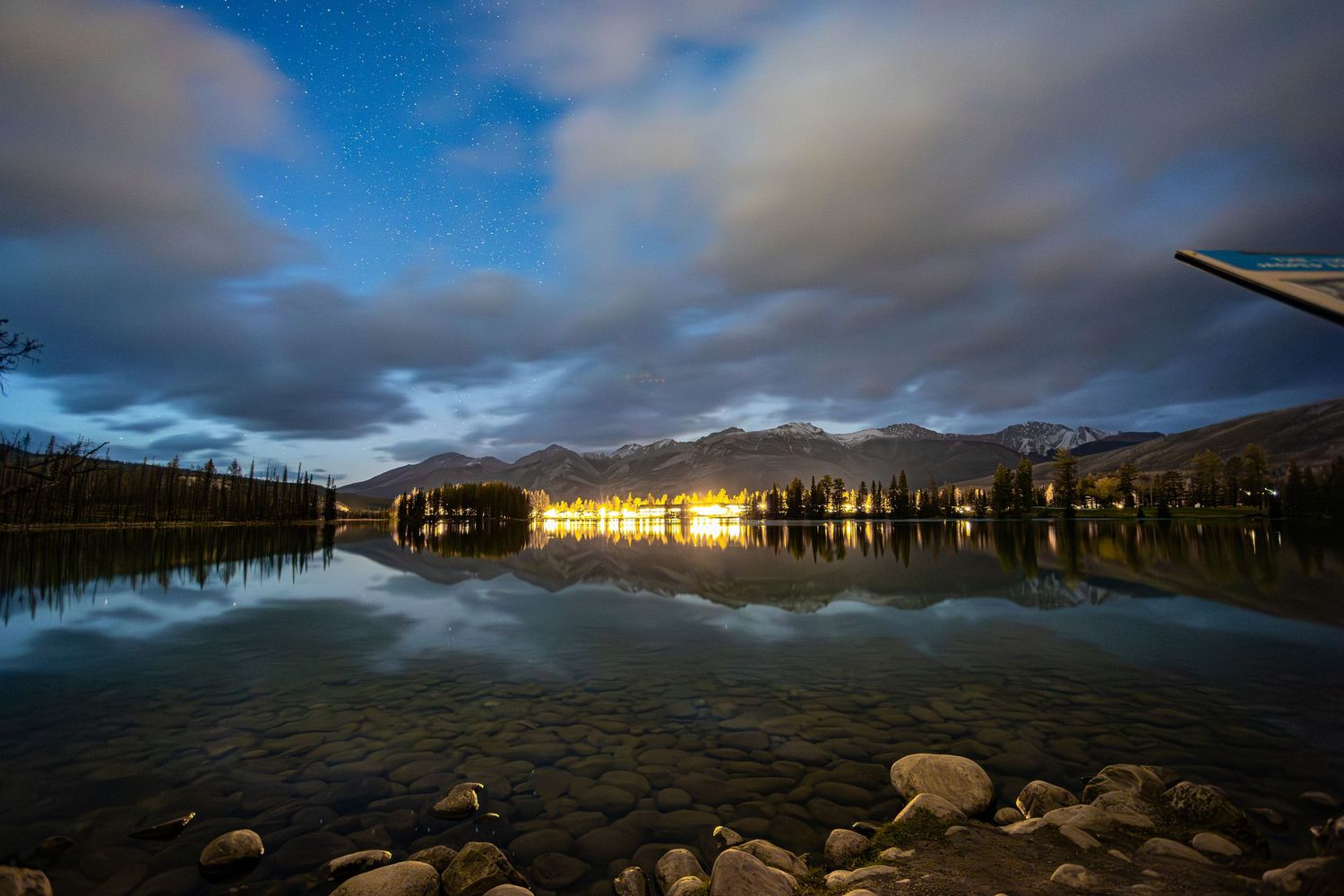 Serene night lake reflecting city lights with mountains and starry sky in the background.