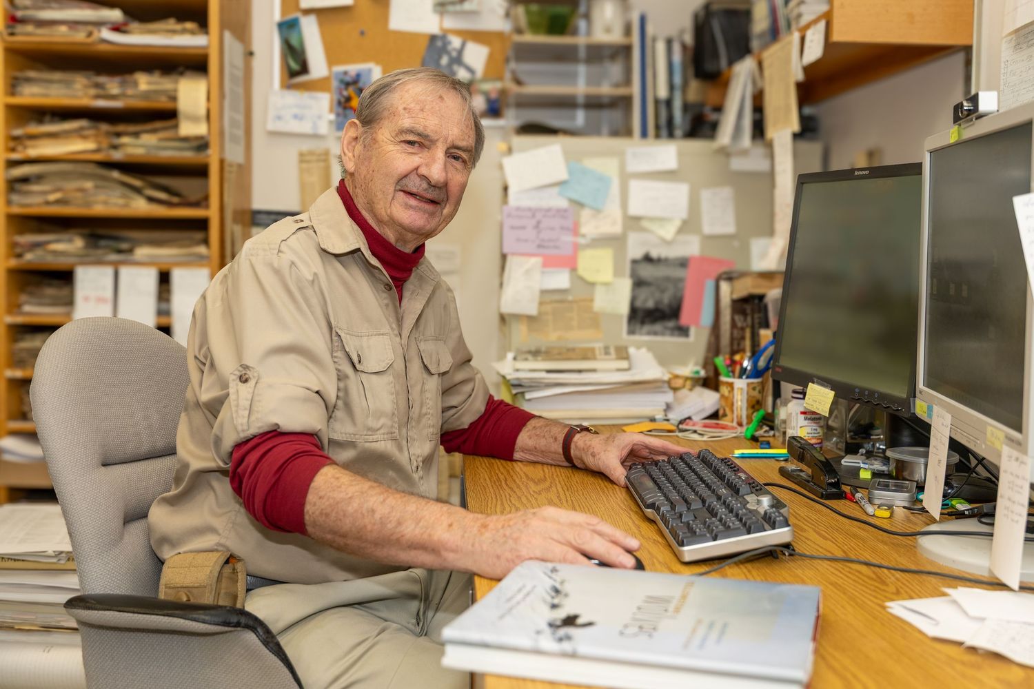 Smiling senior man working at a cluttered office desk with a computer keyboard and monitor.