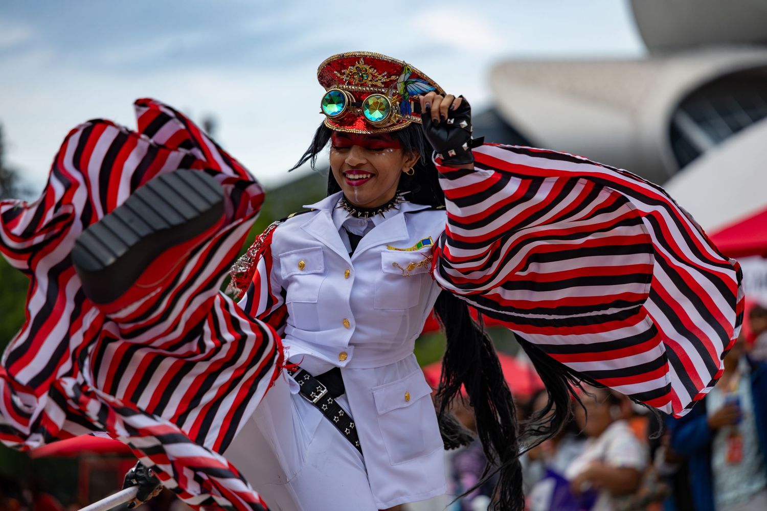 Woman in white military uniform dances with red and white striped fabric at a colorful cultural parade.