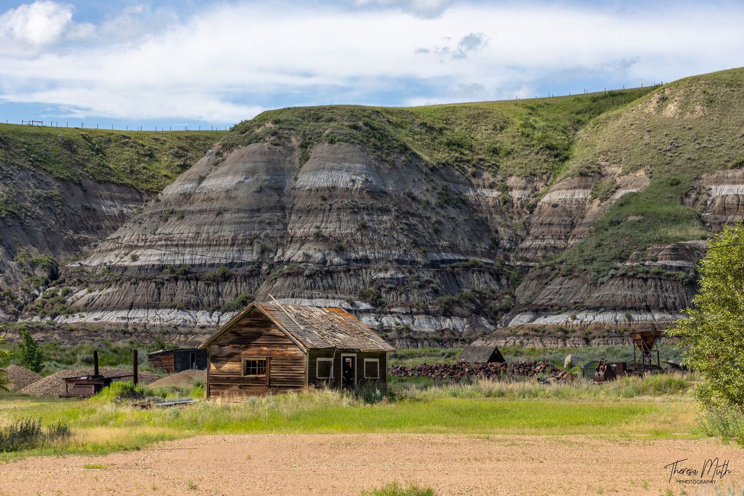 Abandoned wooden homestead buildings nestled against dramatic layered badlands cliffs in a rural Alberta landscape.