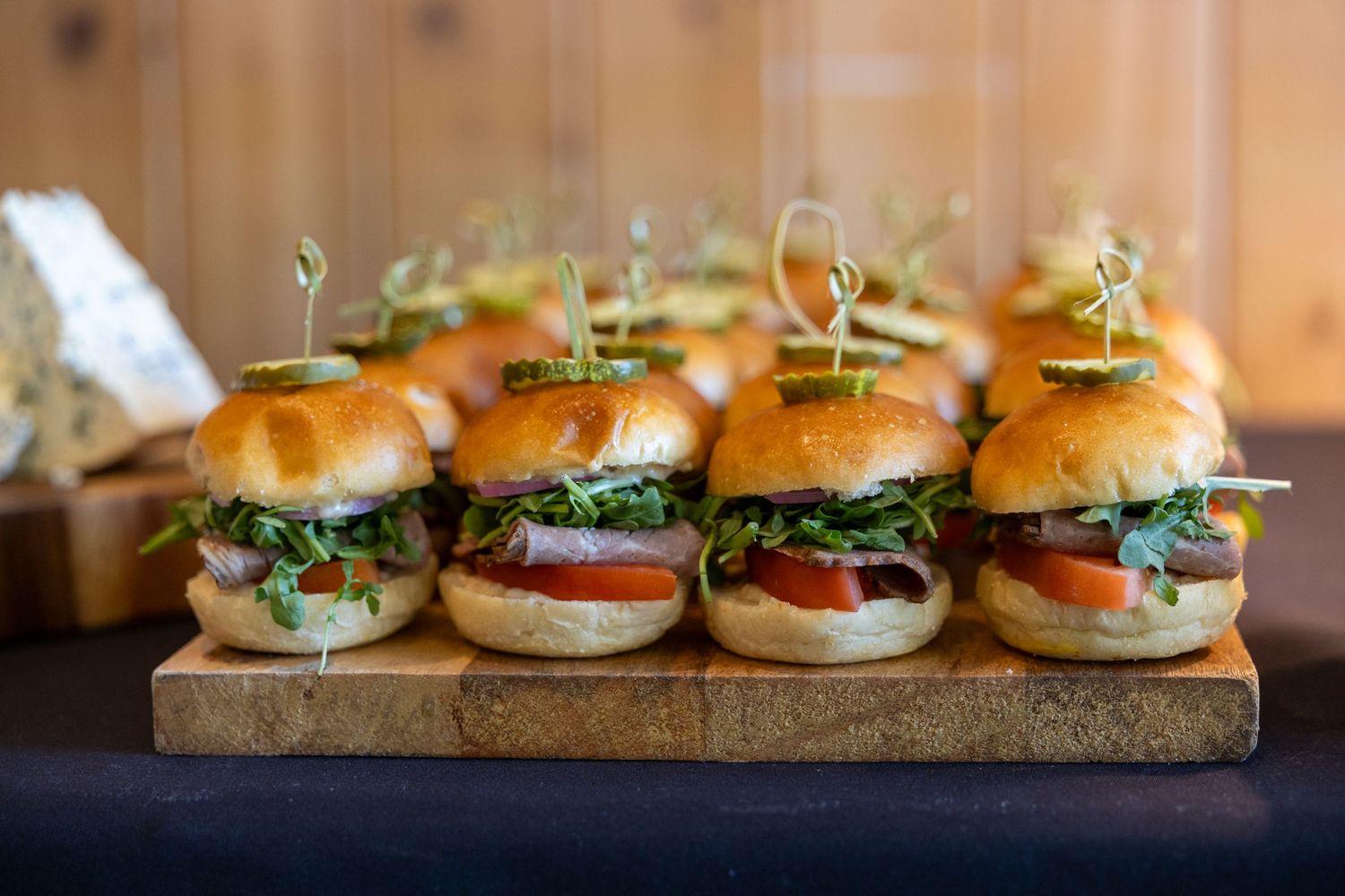 Gourmet mini sliders on a wooden board, topped with fresh greens, tomato, and microgreens at a catered event.