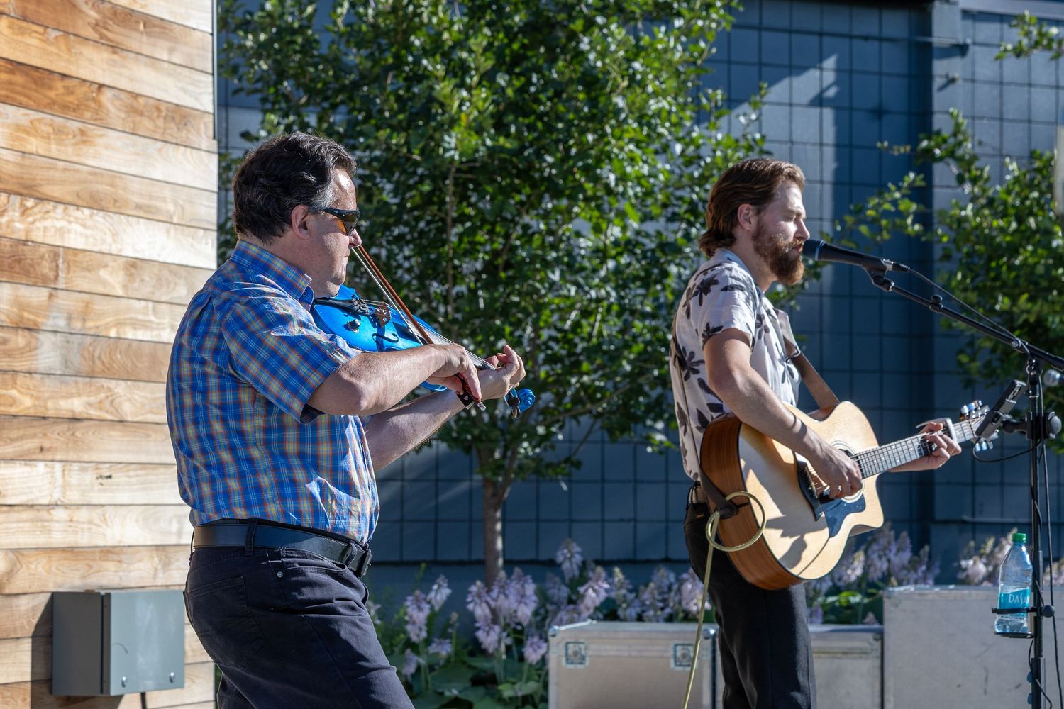 Two male musicians perform outdoors, one playing violin and one playing acoustic guitar at a microphone.