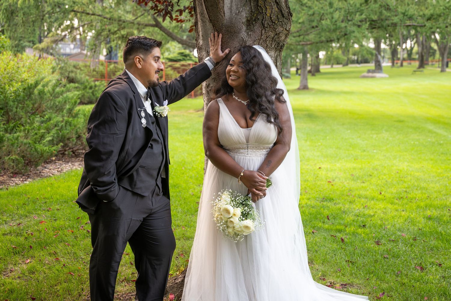 Newlywed couple posing outdoors, groom in black tuxedo touching bride's veil, bride in white gown holding bouquet.