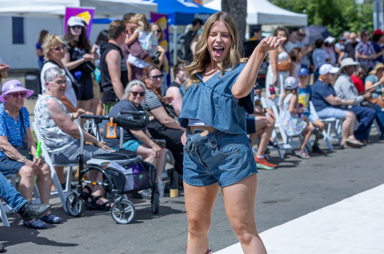 Smiling woman in blue shorts and top energetically walks an outdoor fashion runway before a seated crowd.