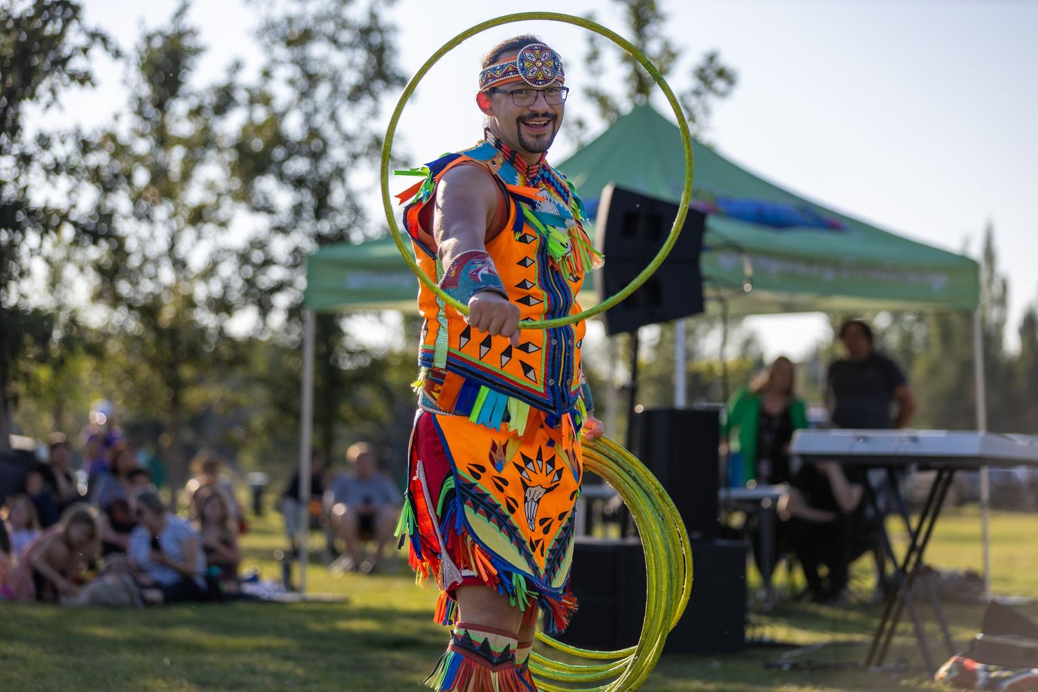 A performer in colorful traditional regalia skillfully spins green hula hoops at an outdoor festival.