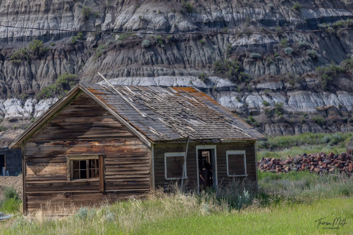 Abandoned wooden homestead cabin in a grassy field with dramatic badlands striped rock formations in the background.