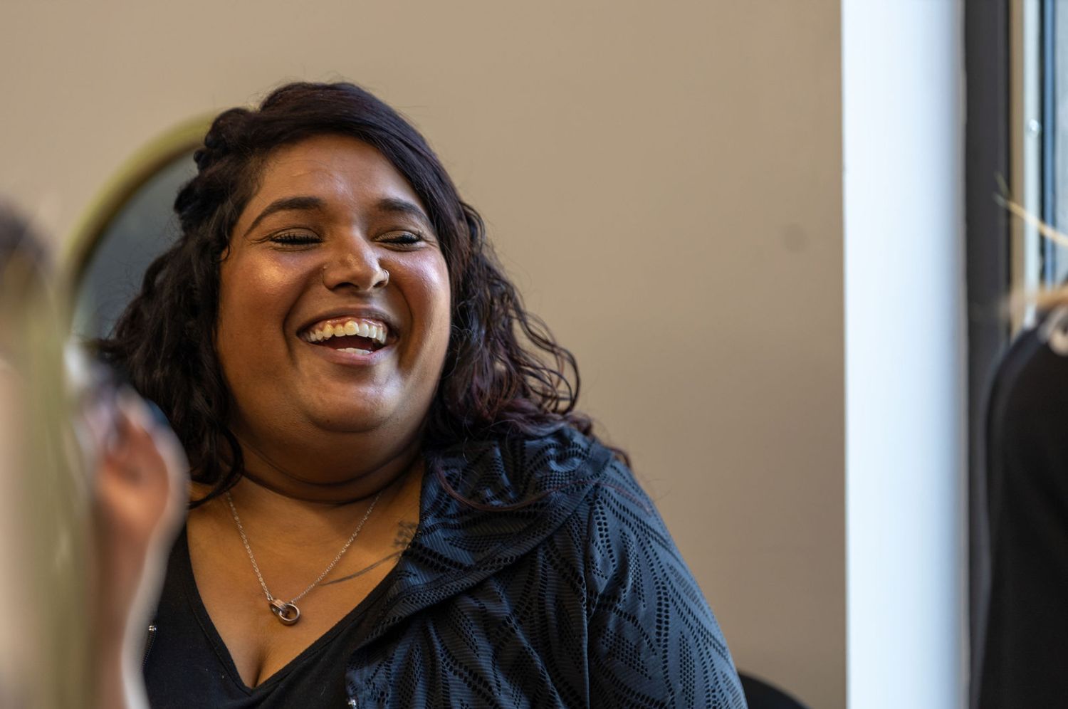 Happy young woman with dark curly hair laughing joyfully near a mirror indoors.