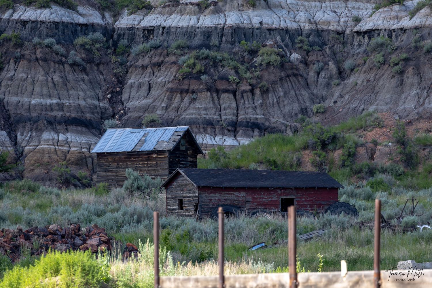 Rustic wooden barns near striped badlands cliffs with sagebrush and a wire fence in the foreground.