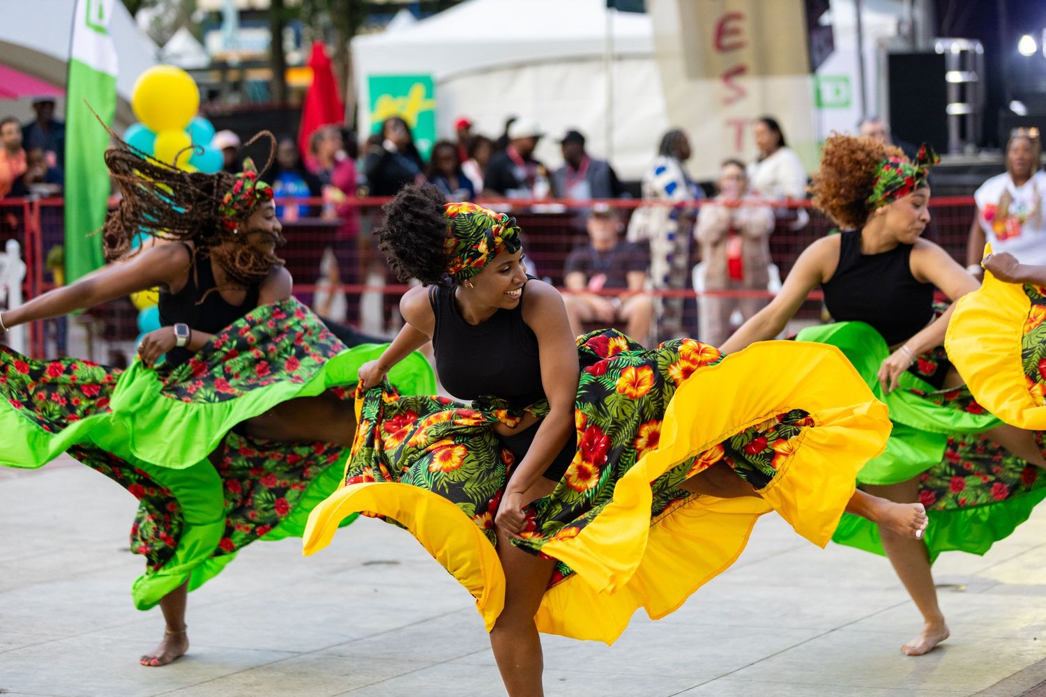 Three women perform Caribbean dance in vibrant floral skirts at an outdoor cultural festival.
