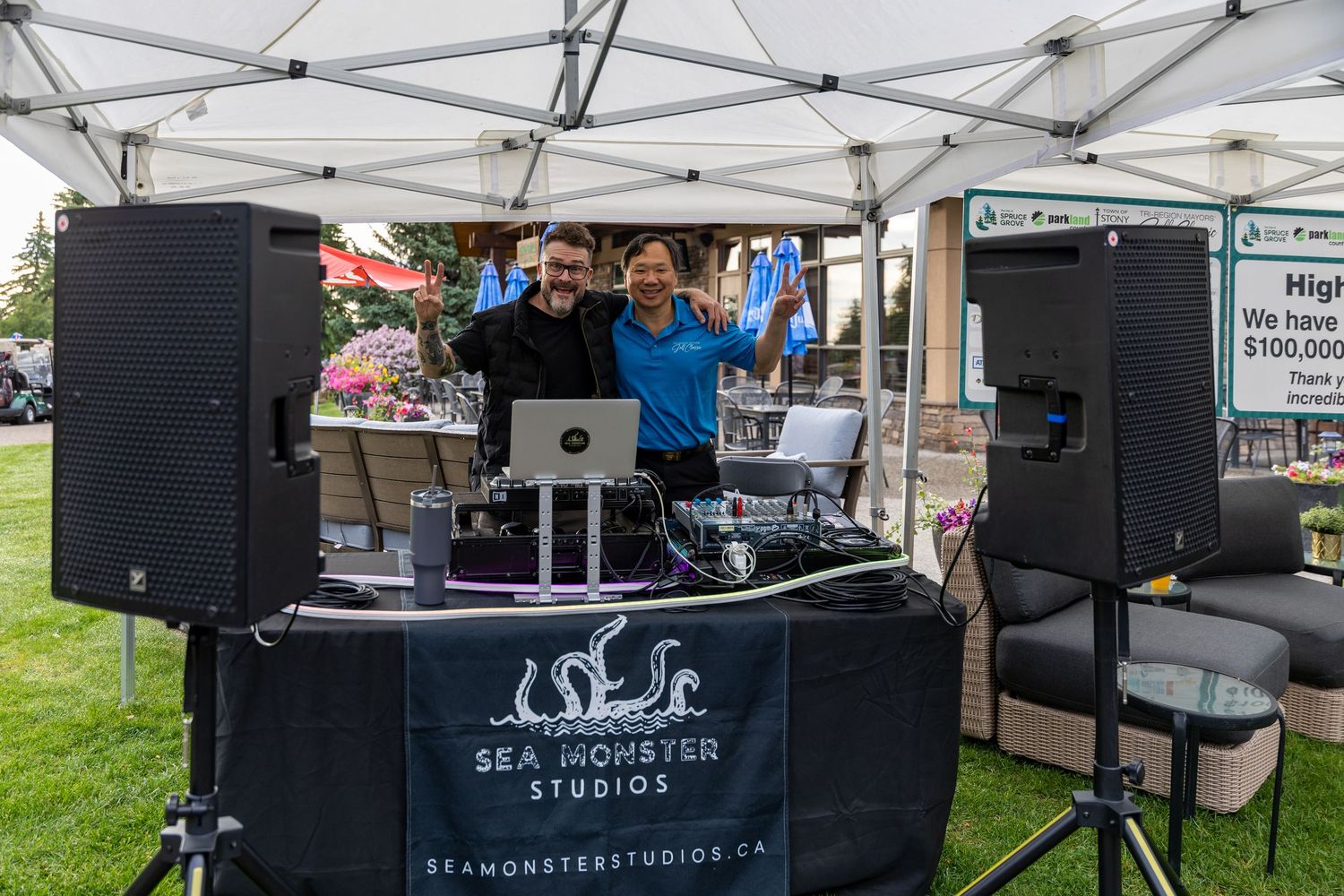 Two DJs pose behind a Sea Monster Studios branded booth with speakers at an outdoor event.