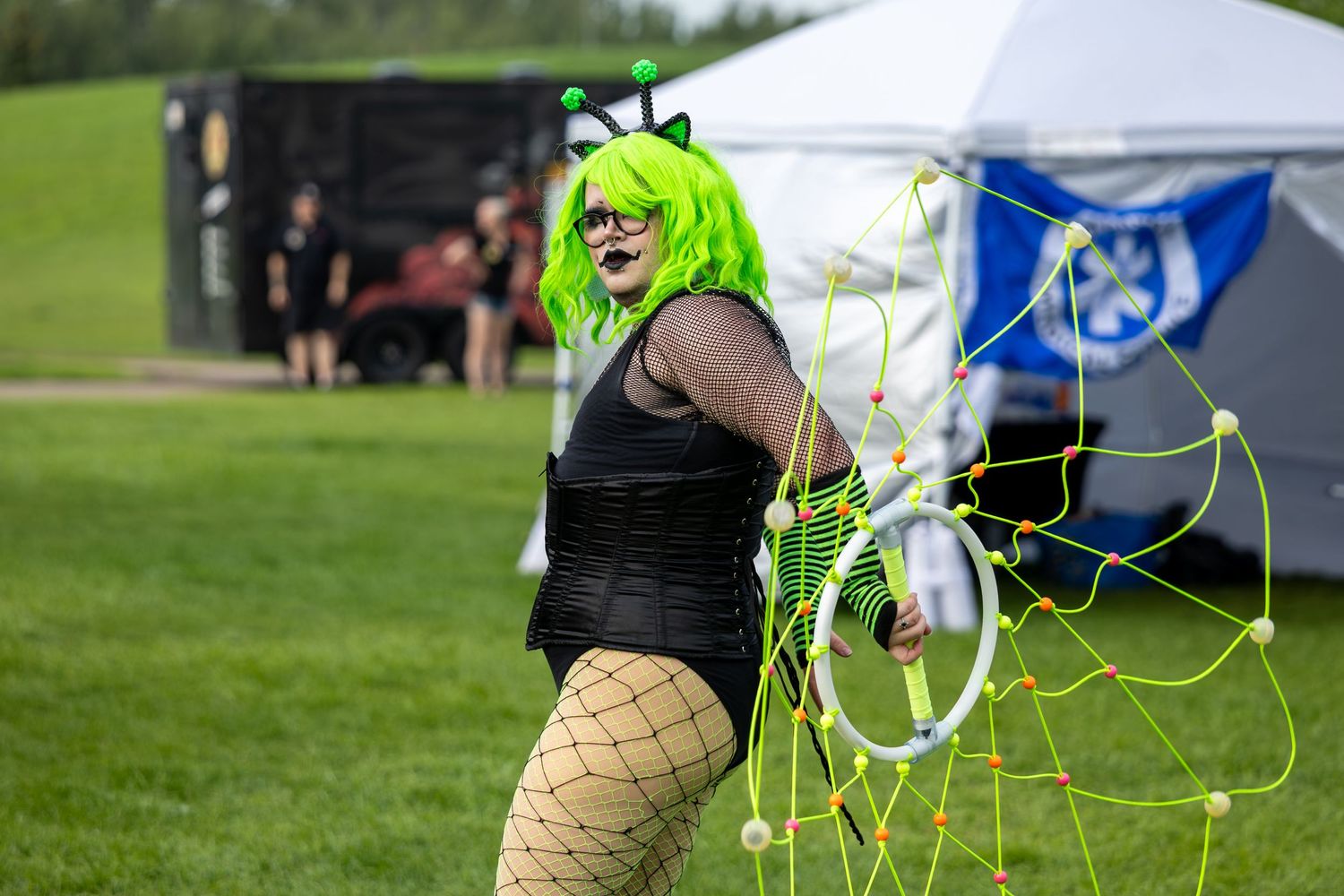 Person in green wig, black outfit, and fishnet stockings holds a neon green web prop at an outdoor festival.