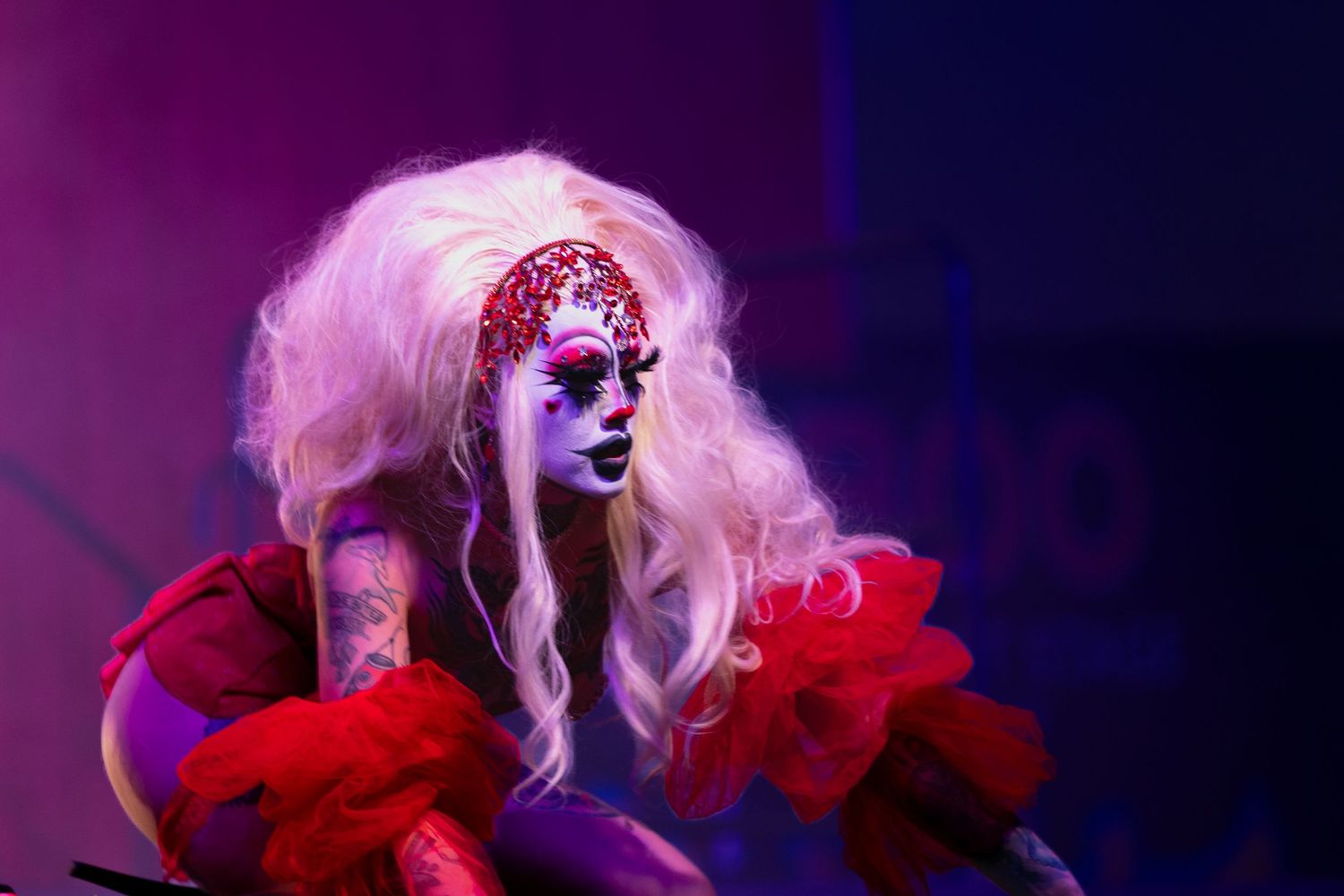 Dramatic drag performer in white wig and red ruffled costume with elaborate face paint under purple stage lighting.